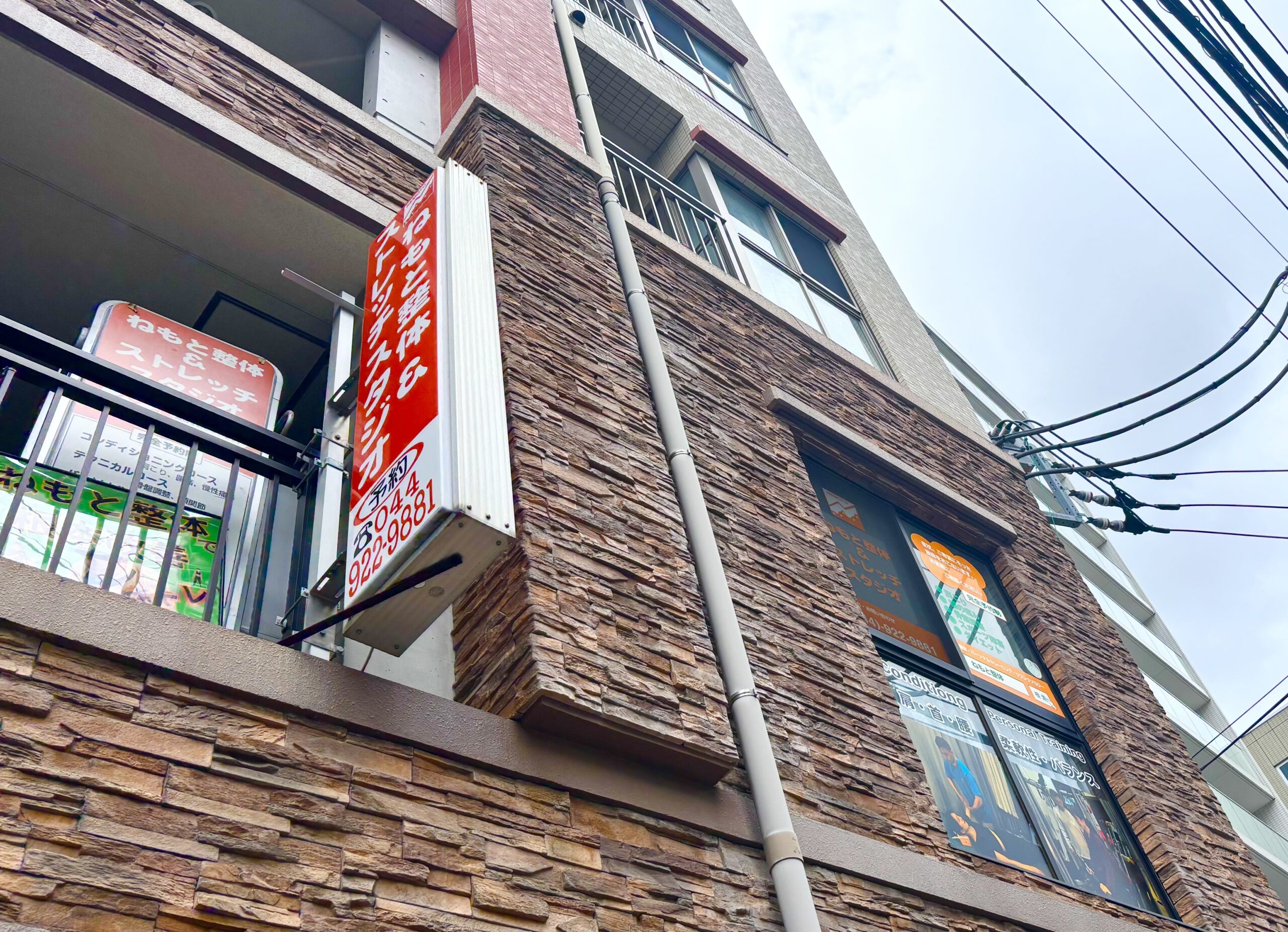 向ヶ丘遊園ねもと整体&ストレッチスタジオの外観。茶色いレンガ調の多階建て建物で、赤い看板と窓に施術案内のポスターが掲示されている