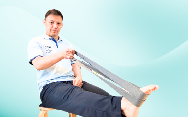 ストレッチバンドを使って足の柔軟性を高める男性整体師。椅子に座りながらふくらはぎとハムストリングを伸ばすストレッチを実演