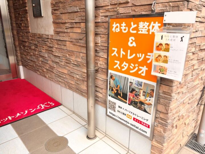 向ヶ丘遊園ねもと整体&ストレッチスタジオの入口。オレンジ色の看板と施術風景の写真が掲載された案内板、赤いマットが敷かれたエントランス
