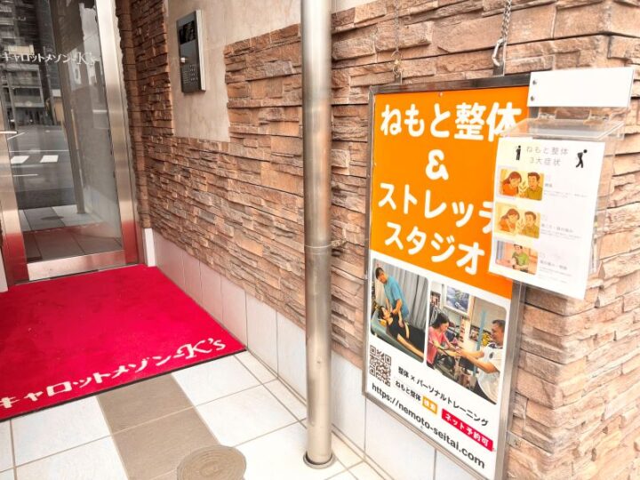登戸ねもと整体&ストレッチスタジオの入口。茶色いレンガ調の壁面にオレンジ色の看板と施術写真の案内板、赤いマットが敷かれたエントランス