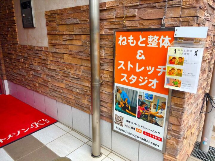 多摩区ねもと整体＆ストレッチスタジオの入口。茶色いレンガ調の壁面にオレンジ色の看板と施術写真の案内板、赤いマットが敷かれたエントランス