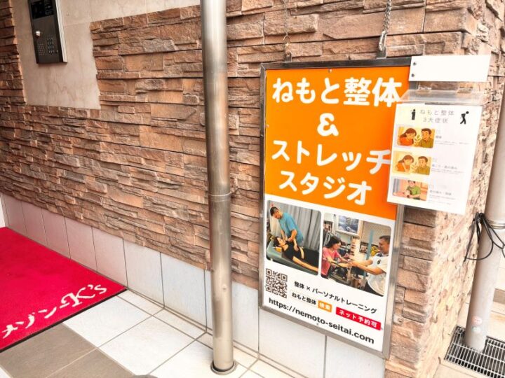 川崎市ねもと整体＆ストレッチスタジオの入口。茶色いレンガ調の壁面にオレンジ色の看板と施術写真の案内板、赤いマットが敷かれたエントランス