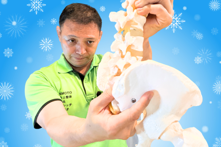 冬の雪景色を背景に腰椎と骨盤の模型を持ち、寒い季節の腰痛対策について解説する院長根本大