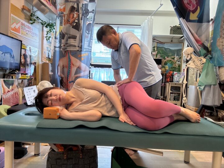 ねもと整体&ストレッチスタジオの院内で、根本院長が女性患者に対して側臥位での腰部・骨盤調整施術を行っている様子。解剖学模型や専門機器が配置された本格的な施術環境。 SEO重視版ALTテキスト 向ヶ丘遊園・登戸のねもと整体&ストレッチスタジオで腰痛・骨盤調整施術を行う根本院長の専門施術風景 施術内容重視版 ねもと整体&ストレッチスタジオで仙腸関節・腰仙関節調整を受ける患者。側臥位での専門的な骨盤調整施術の実施風景。 設備・環境重視版 解剖学模型や専門機器を備えたねもと整体&ストレッチスタジオで、根本院長による個別対応の骨盤調整施術を受ける女性患者 どの用途でご使用になるかをお教えいただければ、さらに最適化したALTテキストを提案いたします。