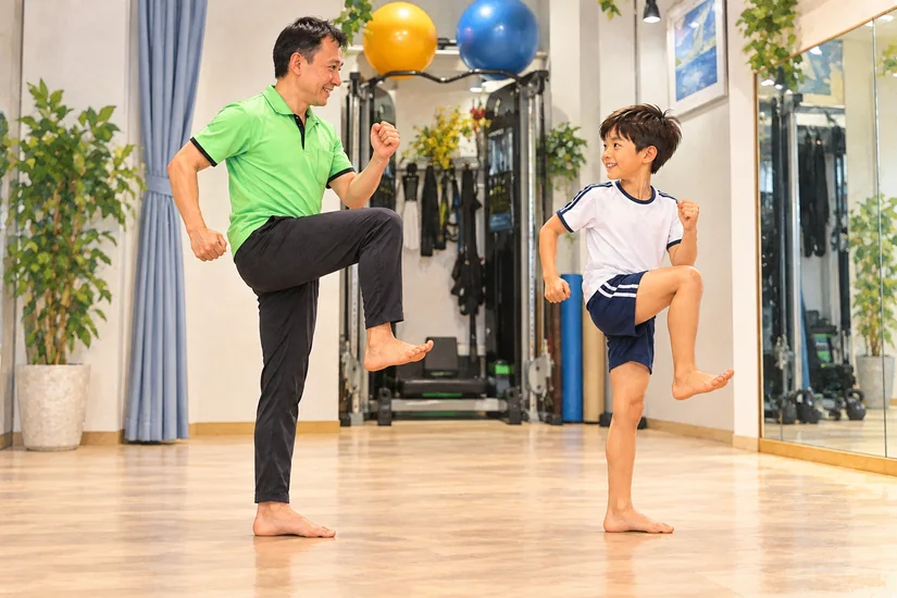 子どもに顔を向けたトレーナーが、裸足で支持脚を伸ばして走りのもも上げを示すイラスト